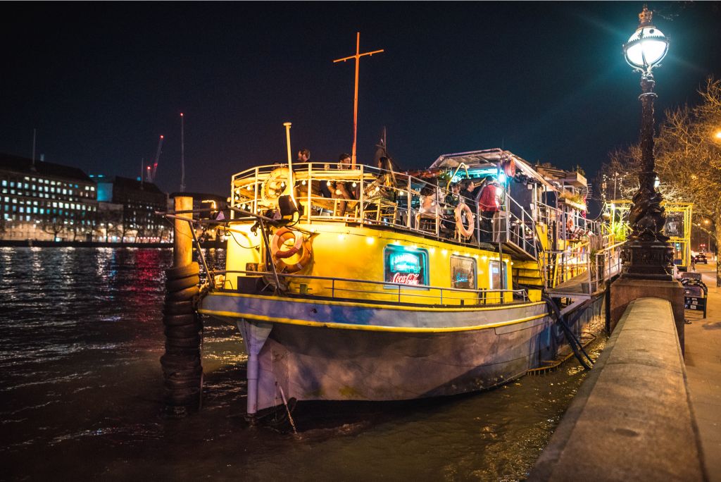 A long barge boat lit up on the Thames with patrons drinking on the rooftop